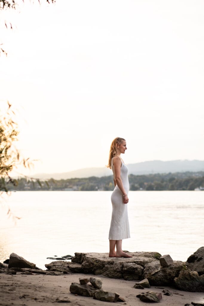 risingqueen- Eine Frau, die ihre persönliche Marke verkörpert, steht barfuß auf einem Felsen am Seeufer und blickt in die Ferne.