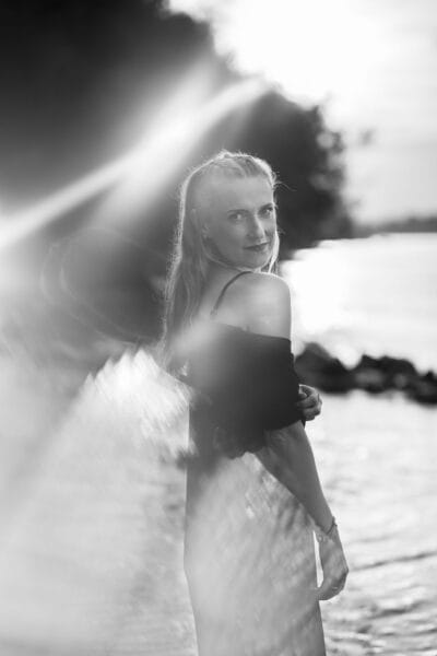 risingqueen- Eine Frau steht am Wasser, während das Sonnenlicht in Schwarz und Weiß aufblitzt und die Essenz des persönlichen Brandings einfängt.