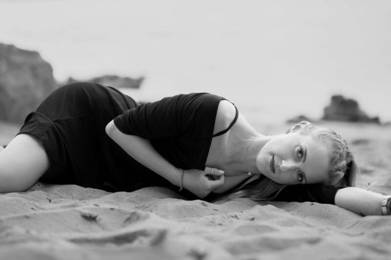 risingqueen- Eine Frau in einem schwarzen Kleid, die ihre persönliche Marke verkörpert, liegt im Sand am Strand.