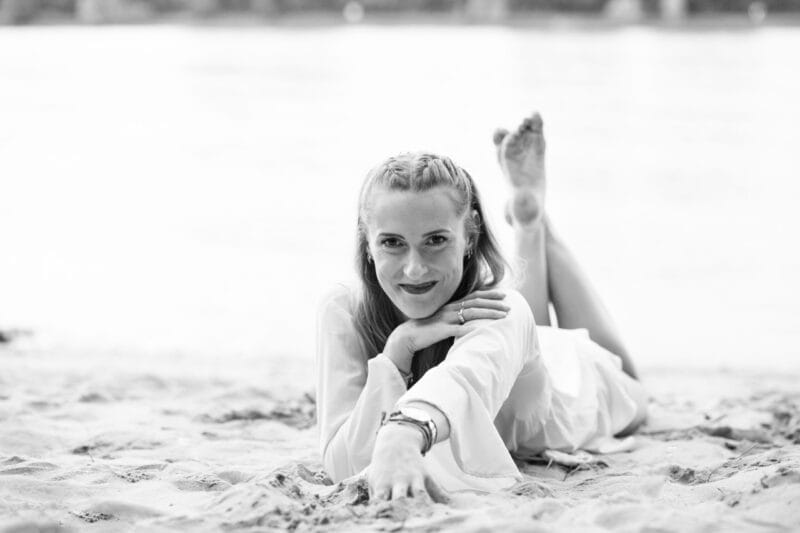 risingqueen- Eine Frau, die in entspannter Pose am Strand liegt, in die Kamera lächelt und das gelassene Branding verkörpert, das wir anstreben.